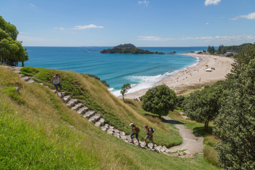 Three people walk up a grassy, stone stairway with a scenic beach in the background. The beach, perfect for the ultimate family getaway, is lined with trees while an expanse of blue ocean, small islands, and a clear sky complete the picturesque view. Swimmers and sunbathers are visible on the beach.