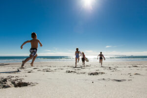 Four children are running towards the ocean on a sunny beach with clear blue skies in Papamoa. The kids, dressed in swimwear, leave footprints in the sand as they race to the water's edge. The sun shines brightly, casting a warm and vibrant light over this perfect day of fun—one of many things to do in Papamoa.