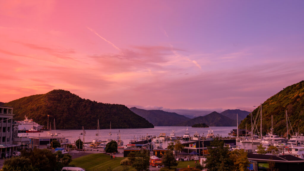 A picturesque marina at sunset with boats docked along the shore, surrounded by lush green hills. The sky is a blend of orange, pink, and purple hues, reflecting on the calm water. Buildings and trees are visible in the foreground, offering a serene view perfect for guests at Tasman Holiday Parks - Picton.