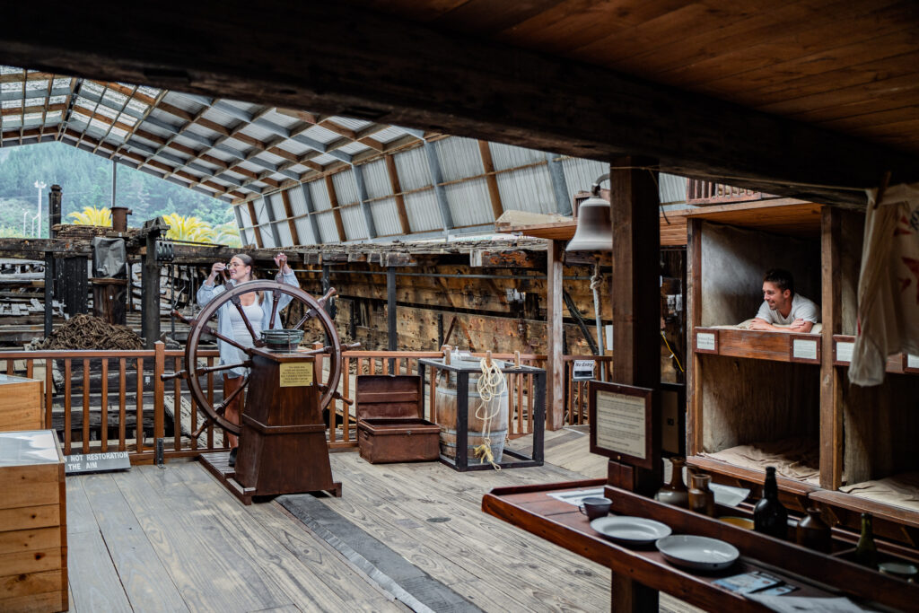 A historical museum exhibit depicting an old ship's interior is among the top things to do in Papamoa. The room features a large wooden ship wheel, various navigation instruments, display cases, and informational placards. Visitors are seen exploring the exhibit, and a man is standing in a lookout box.