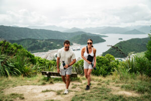 A man and a woman hike up a grassy trail with lush green hills and a sprawling body of water visible in the background, capturing the essence of Tasman Holiday Parks - Picton. The man holds a water bottle, and both wear casual athletic attire and sunglasses. A wooden bench is set along the trail. A recommendation in the ultimate guide to Picton.