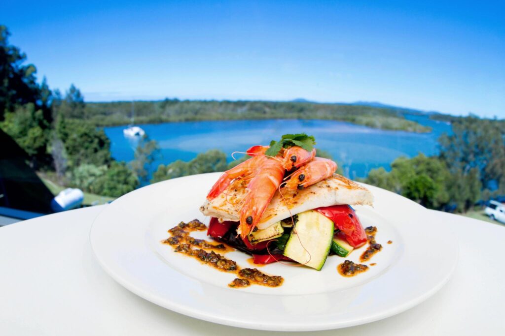 A gourmet dish featuring grilled fish topped with shrimp and assorted vegetables, garnished with a drizzle of sauce, is presented on a white plate. The background displays a serene lake scene with trees and a boat under a clear blue sky.