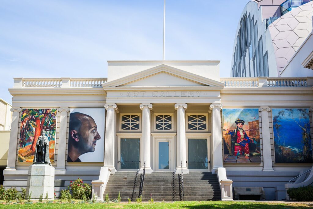 The facade of a classical-style art gallery with three grand doors and columns stands impressively, perfect for a road trip stop. Two large, colorful paintings adorn either side of the entrance. A small statue graces the left side of the steps, with a modern building peeking from behind.