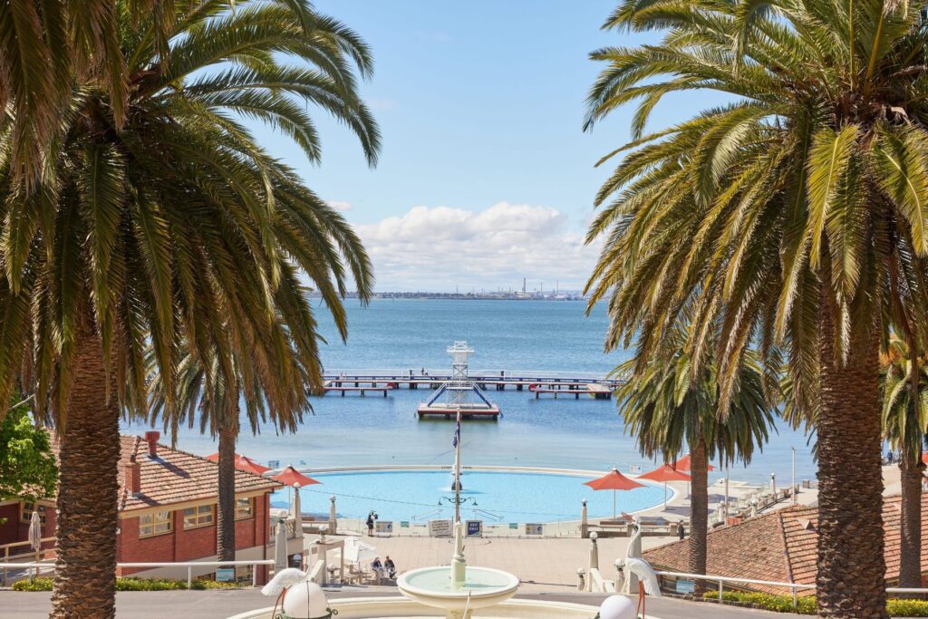 A scenic view features a large fountain in the foreground, bordered by tall palm trees. Steps lead down to a pool area with red umbrellas and seating. In the background, a pier extends into a calm, blue body of water under a clear sky with scattered clouds—perfect for your road trip stopover.