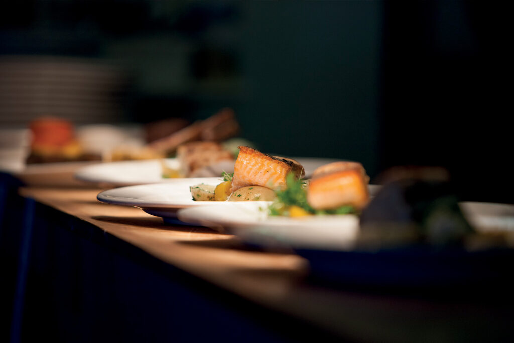 Three white plates on a wooden table, each showcasing a piece of cooked salmon alongside roasted vegetables. The background is blurry, bringing attention to the plated food in the foreground—one of the many delightful things to do in Papamoa.