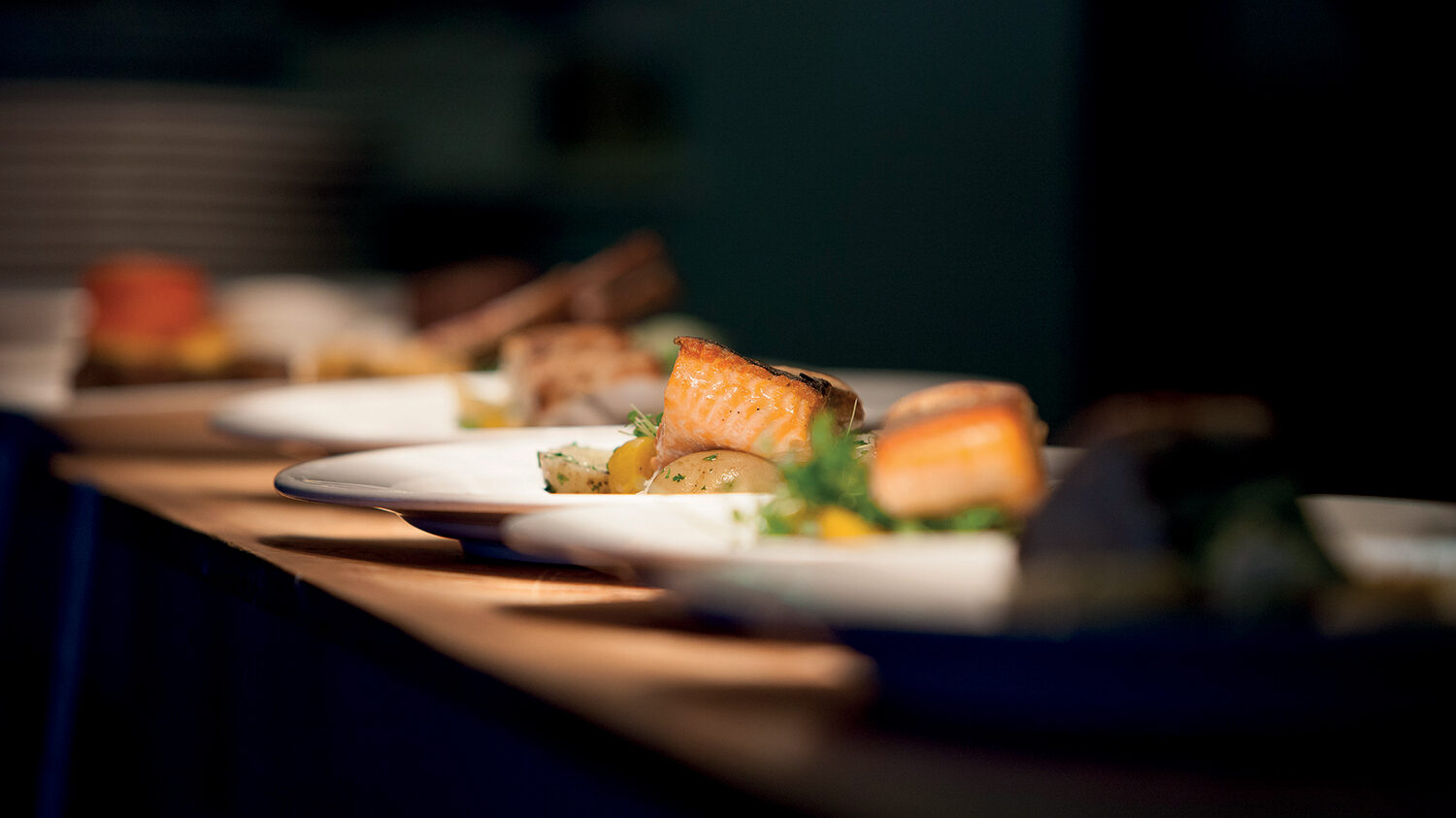 Three white plates on a wooden table, each showcasing a piece of cooked salmon alongside roasted vegetables. The background is blurry, bringing attention to the plated food in the foreground—one of the many delightful things to do in Papamoa, found in our Ultimate guide to Papamoa. 