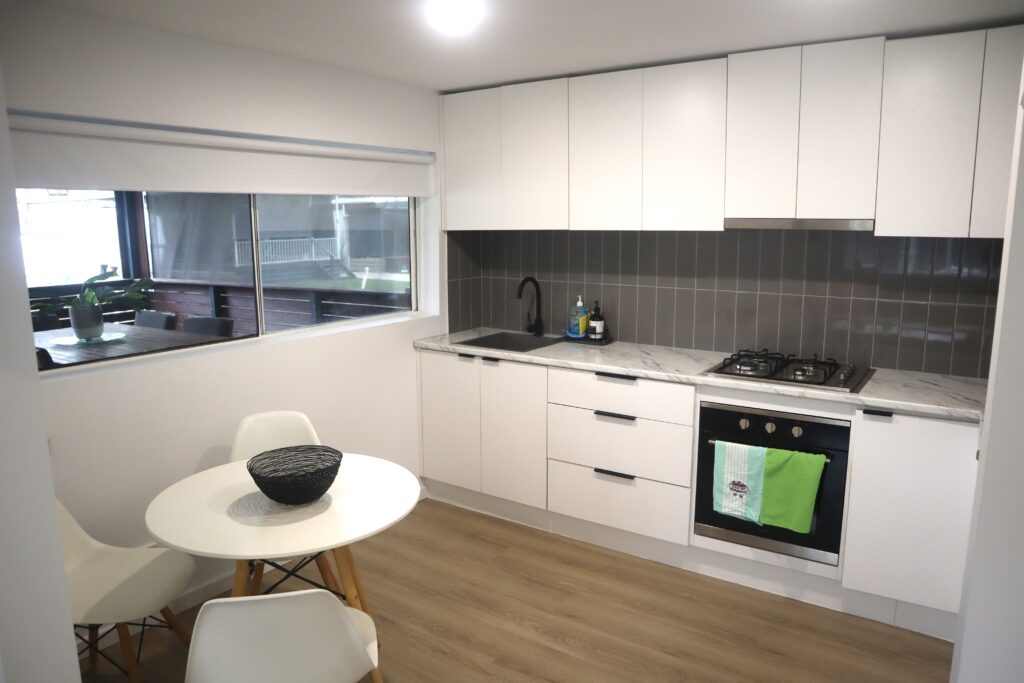 A modern kitchen with white cabinets and drawers, a gray tile backsplash, and a stainless steel gas stove. There is a small round dining table with white chairs and a black bowl centerpiece. A large window provides natural light, and light wood flooring complements the space.