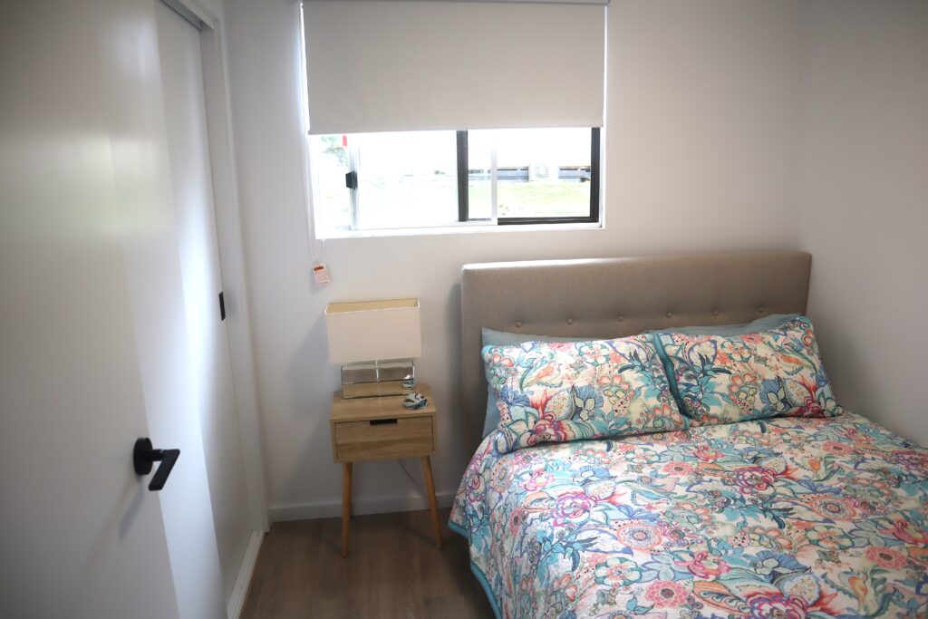 A small bedroom features a bed with a floral-patterned bedspread, a gray tufted headboard, and a bedside table with a lamp and small items. The room has white walls, a window with a roller blind, and a closed door with a black handle.