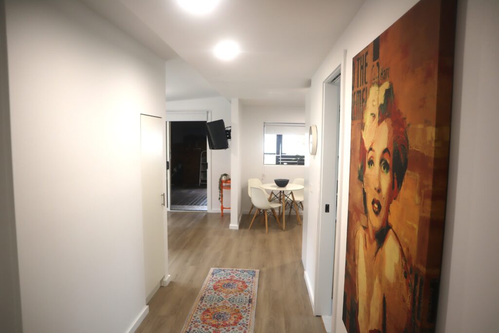 A hallway with wooden flooring leading to a dining area with a white table and chairs. The right wall features a large colorful painting of a woman's face. A small rug with a patterned design lies on the floor. The space is well-lit with ceiling lights.