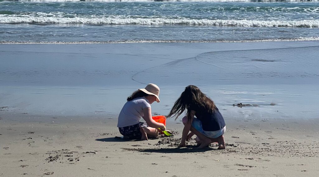 Two girls are sitting on a sandy beach in Papamoa, playing with a red bucket and toys. One girl is wearing a hat and facing the ocean, while the other has long hair and is leaning forward. Waves can be seen hitting the shore in the background—just one of many relaxing things to do in Papamoa.