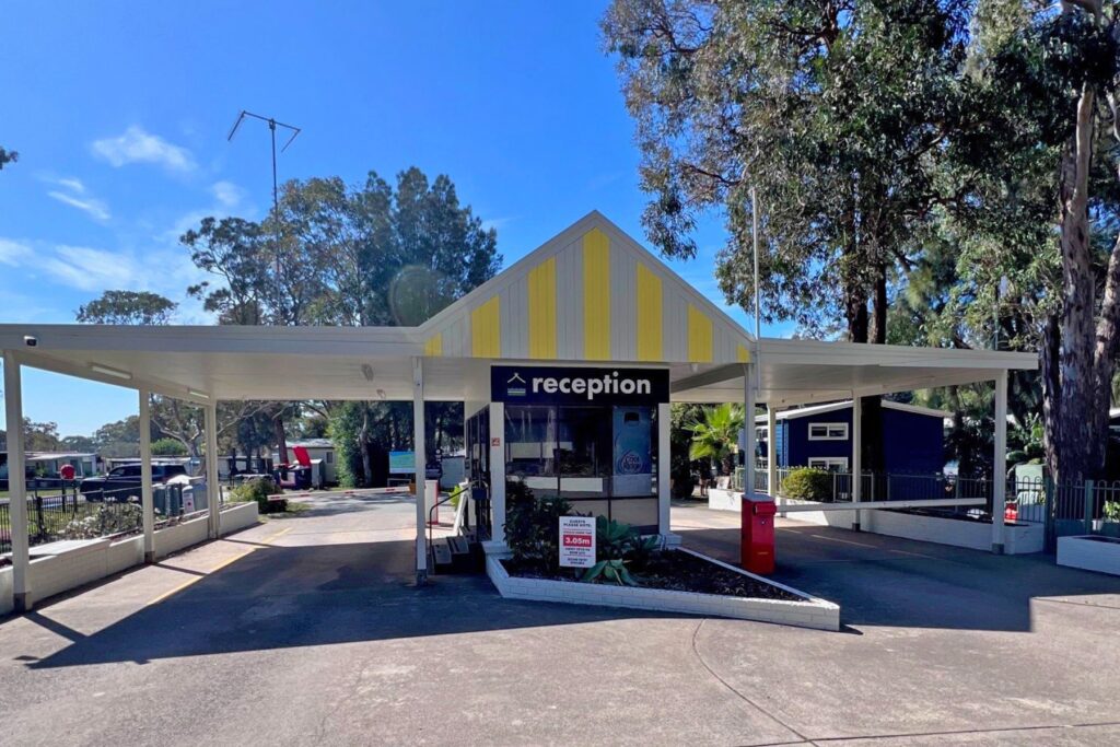 A reception area with a yellow and white striped building, surrounded by trees near Kioloa Beach. The structure has a large covered entrance area and a reception sign above the door. Parking spaces and some vehicles are visible around the building. A clear sky is in the background.