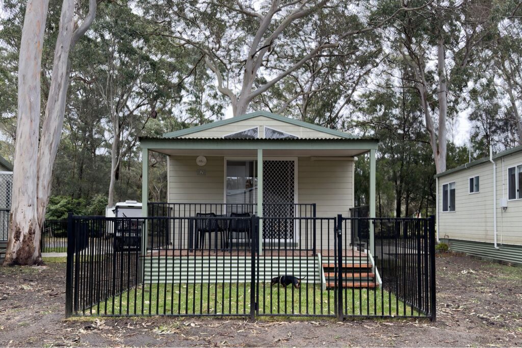 Nestled near Kioloa Beach, this small, light-colored cabin with a green roof is surrounded by tall trees. It features a fenced porch with outdoor furniture, and one step leads up to the entrance where a black dog lounges on the grass inside the fence.