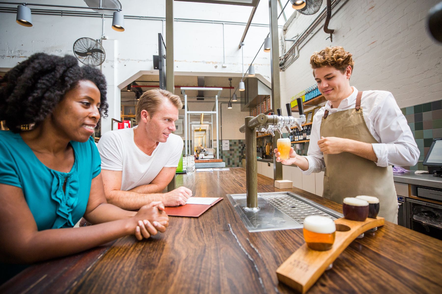 A bartender pours a drink from a tap for two patrons at a wooden bar, reminiscent of a cozy stop on a road trip. The bartender wears a white shirt and beige apron, while the patrons, a woman in a blue blouse and a man in a white t-shirt, smile and engage in conversation. Sample drinks on a wooden tray are in the foreground.