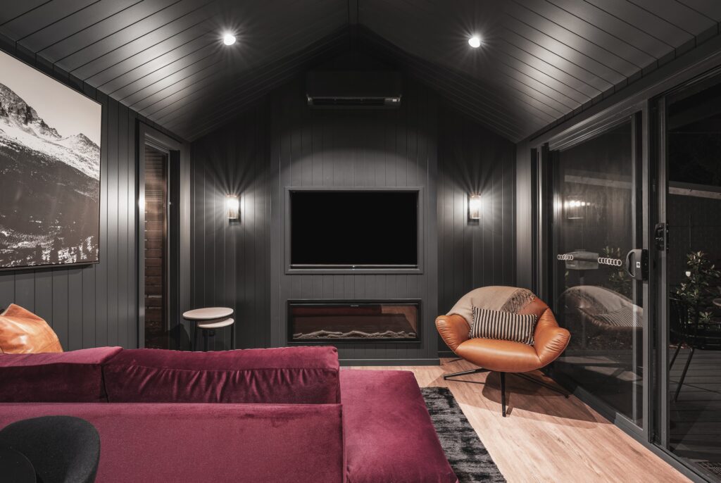 A cozy, modern living room with bright touches against dark walls and ceiling, featuring a flat-screen TV, a fireplace, and a red sofa. A round chair with a striped pillow sits by glass sliding doors. A large black and white mountain photo decorates the wall.