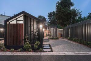 Modern black house with angular roof, featuring bright large windows and a minimalist design. The exterior is accentuated with wooden slats and a small landscaped garden. A pathway leads to the entrance, flanked by greenery under a twilight sky.