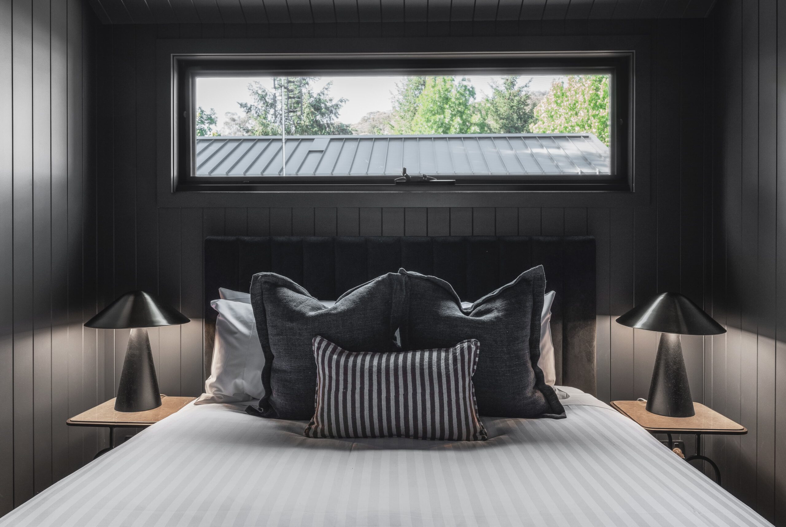 A modern bedroom with dark walls features a neatly made bed with a bright, striped white cover and several black and gray pillows. Two black lamps rest on wooden bedside tables beside the bed. A rectangular window provides natural light.