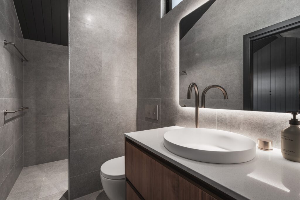 Modern bathroom with bright gray tiled walls and floor. Features a round white sink on a wooden vanity, sleek faucet, and a large mirror with backlighting. A walk-in shower with a glass partition is on the left. Soap dispenser and candle add warmth to the counter.