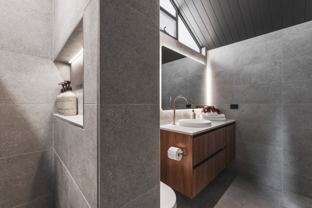 Modern bathroom with gray tiled walls and floor, featuring a wooden vanity with a bright white sink and circular mirror. A soap dispenser sits on an inset shelf. Recessed lighting provides a soft glow, while a window above bathes the space in natural light.