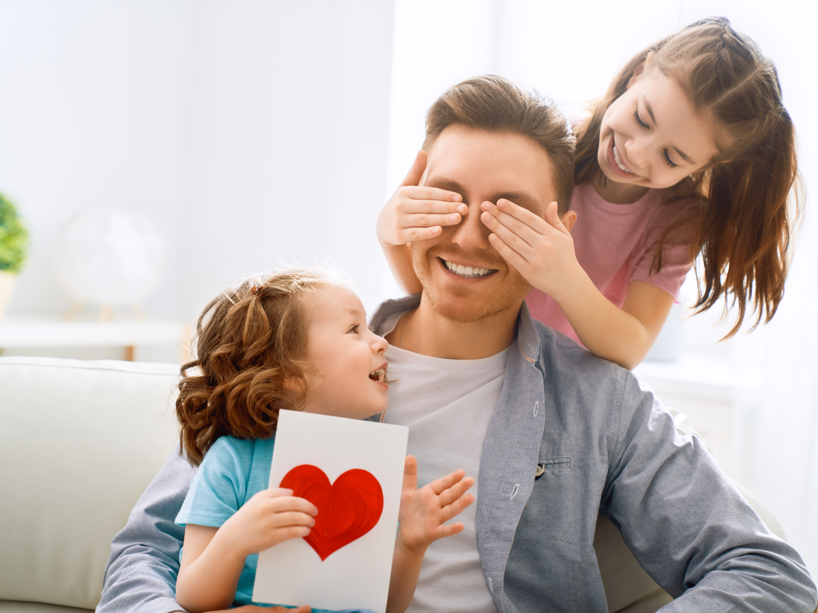 A man is sitting on a couch, smiling, while a young girl covers his eyes and another young child holds a card with a large red heart in front of him. They are all laughing and appear to be enjoying a playful moment together after coming back from the local sausage sizzle.