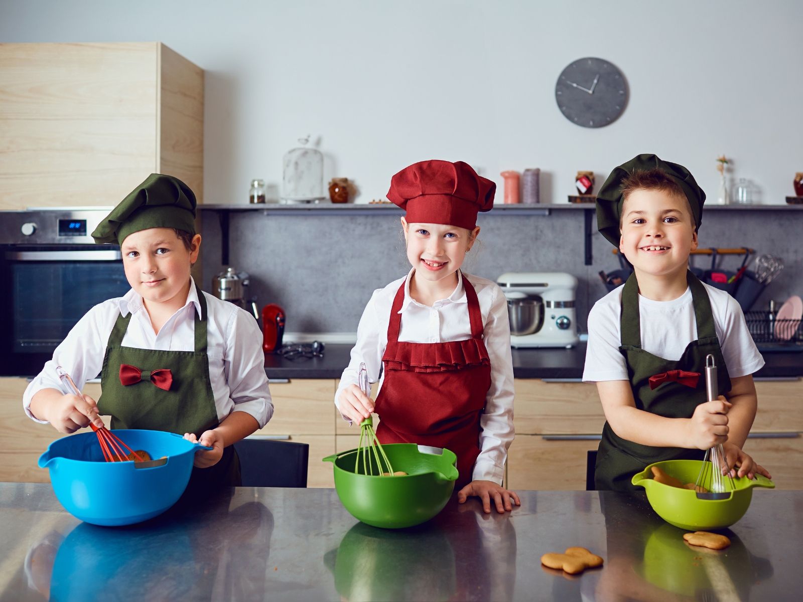 Three children wearing chef hats and aprons are baking in a kitchen. Each child is holding a whisk and mixing ingredients in a colorfully distinct bowl—blue, green, and yellow. They stand in front of a counter with various utensils and a wall clock in the background.