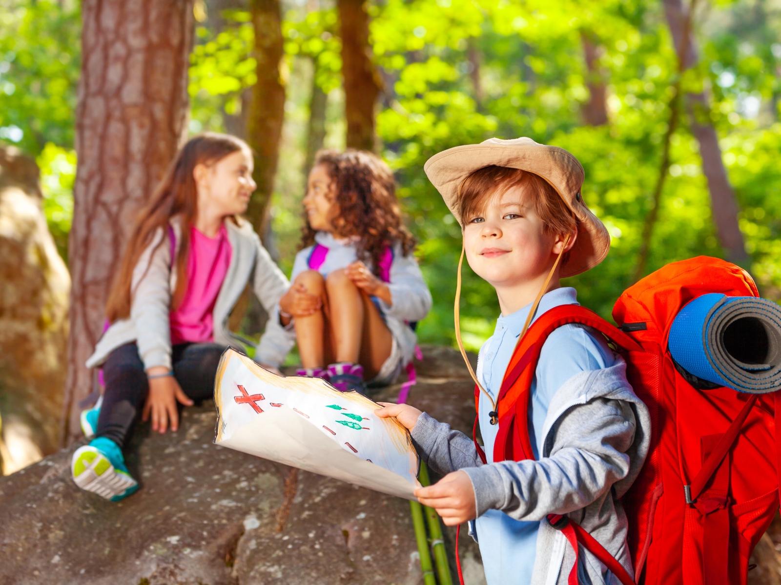 A smiling child stands in the foreground holding a map and wearing a sun hat and a large red backpack. In the background, two children sit on a rock, chatting and smiling, surrounded by lush green trees. The scene depicts an outdoor adventure or camping trip.