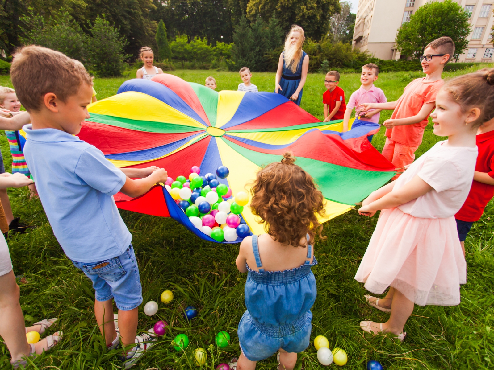 A group of children is holding a colorful parachute in a grassy field, lifting it high. Some plastic balls are scattered on and around the parachute. Trees and buildings are in the background, and a few adults are supervising the activity.