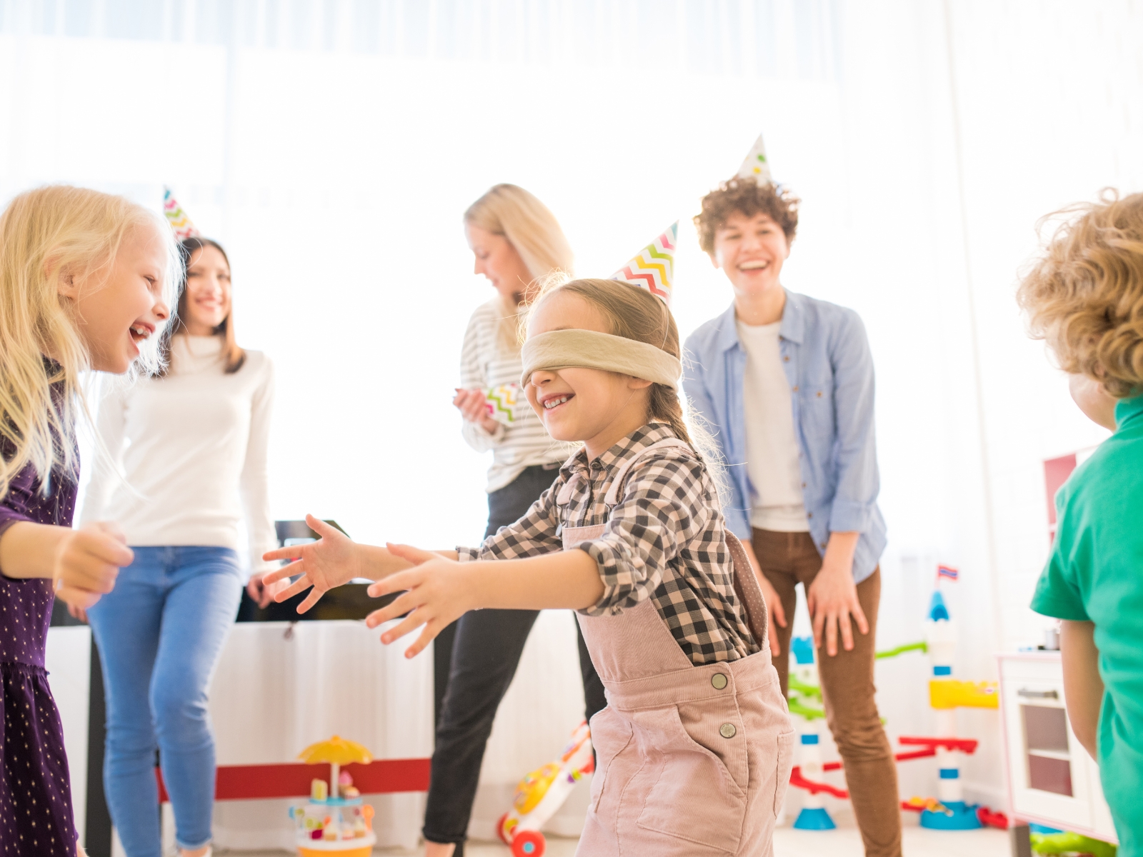 A group of children and adults at a party are playing a game. One child is blindfolded in the center, reaching out to find others, while the surrounding kids laugh and try to avoid being tagged. Everyone is wearing party hats and enjoying the activity.