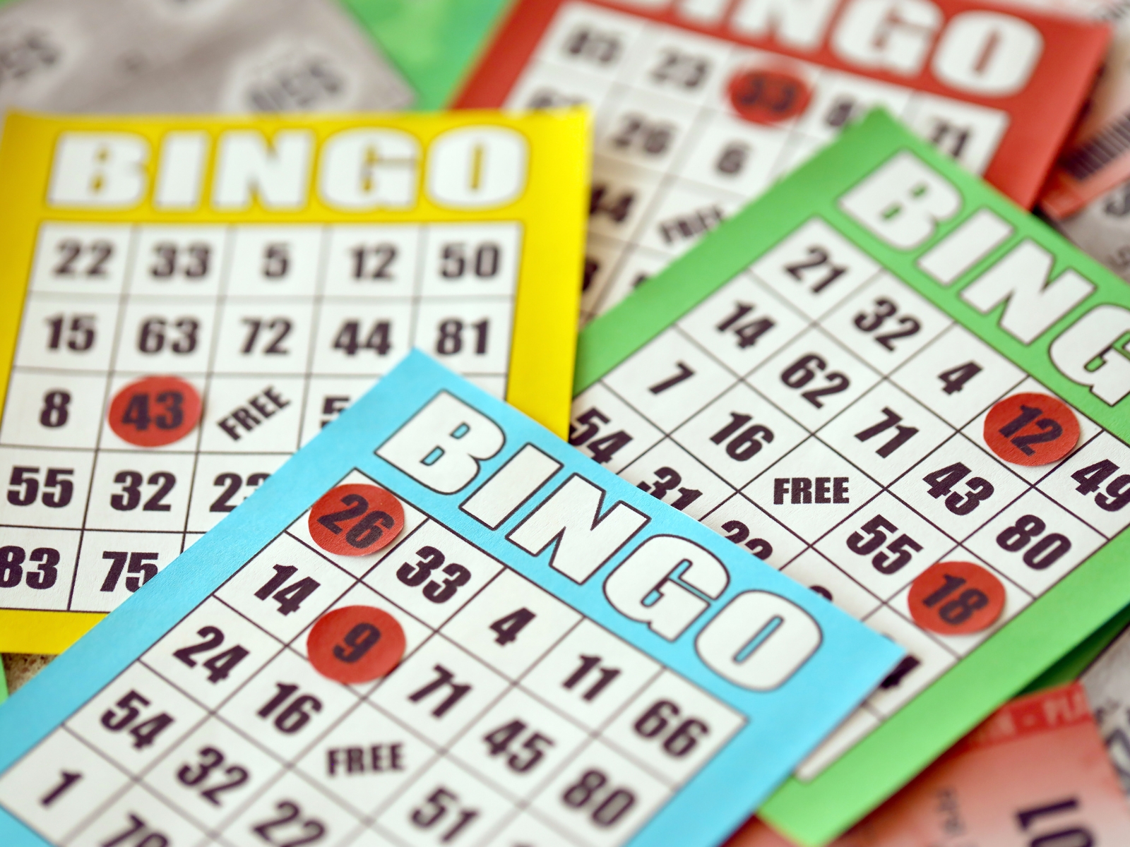 Close-up of multiple bingo cards in various colors, including yellow, green, and blue, scattered on a surface. The cards display a mix of numbers and some numbers are marked with red circles, indicating they have been called.