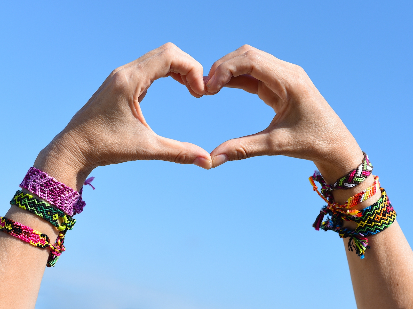 Two hands forming a heart shape against a clear blue sky, adorned with colorful woven friendship bracelets on both wrists. The intricate patterns and bright colors of the bracelets stand out prominently.
