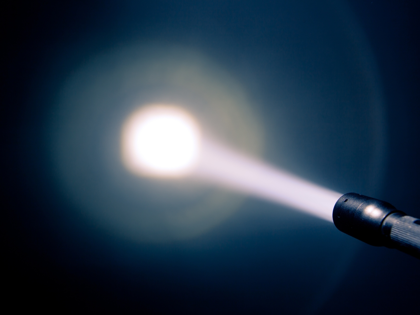 A focused beam of bright white light shines from the tip of a flashlight against a dark background, creating a circular halo effect around the central light beam. The light source is directed outward, cutting through the surrounding darkness.