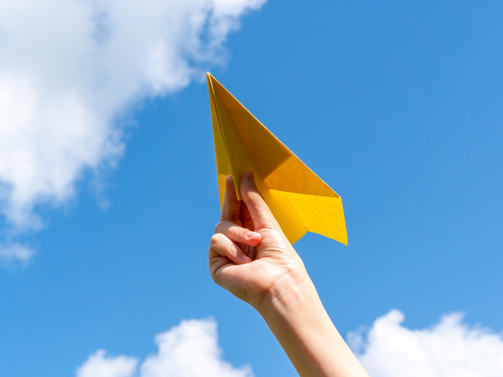 A hand holding a yellow paper airplane up against a bright blue sky with scattered white clouds. The airplane is in sharp focus, capturing a sense of whimsy and freedom.