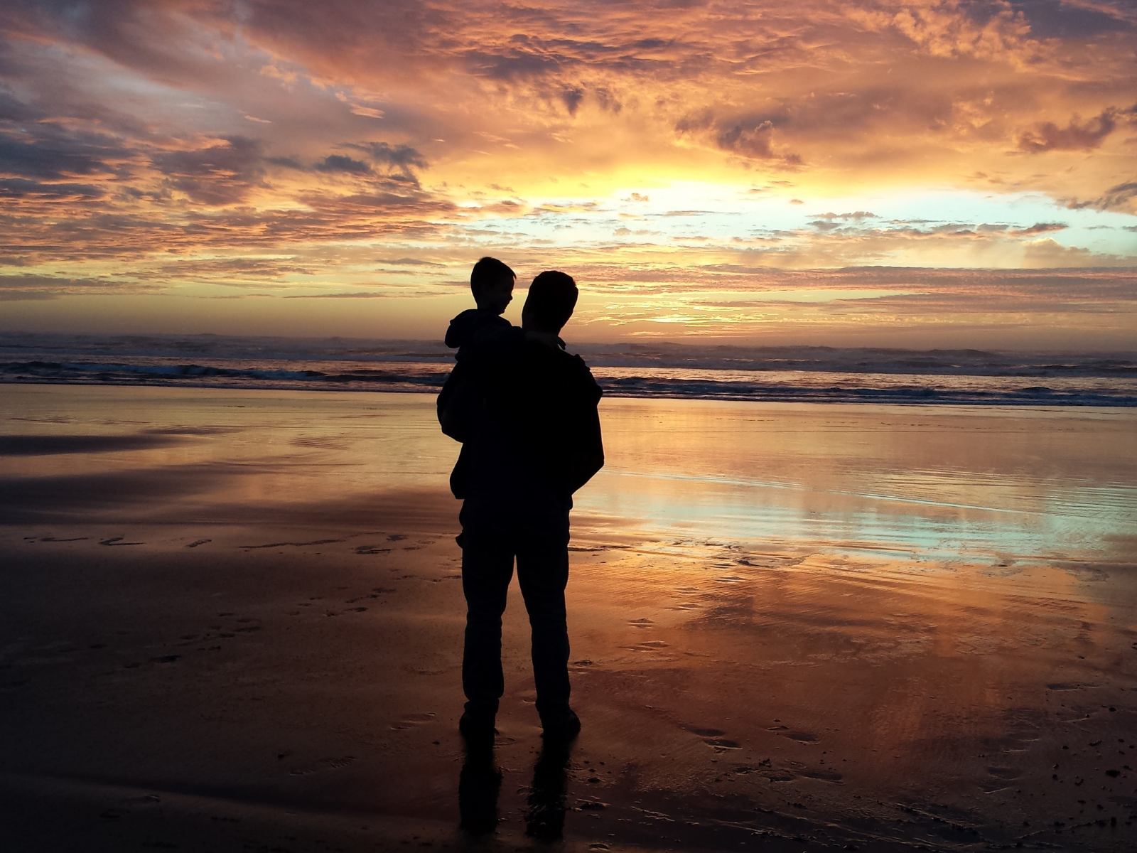 A person holds a child while standing on a beach, facing the ocean at sunset. The sky is filled with vibrant hues of orange, pink, and purple, reflected on the wet sand. The tranquil scene captures a moment of bonding against a stunning natural backdrop.