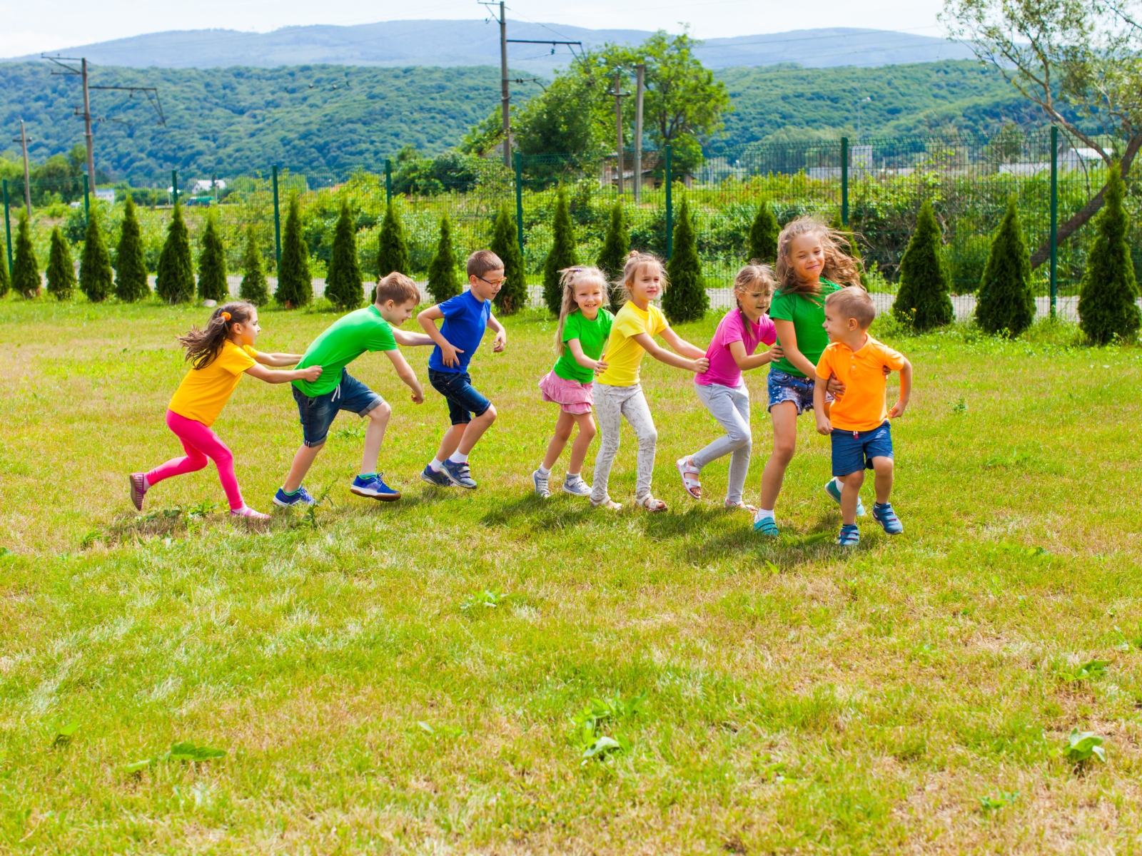 A group of children, dressed in colorful shirts and shorts, play a game in a grassy field with distant green hills. They form a line holding each other’s shoulders, smiling and laughing, with trees and a fence in the background.