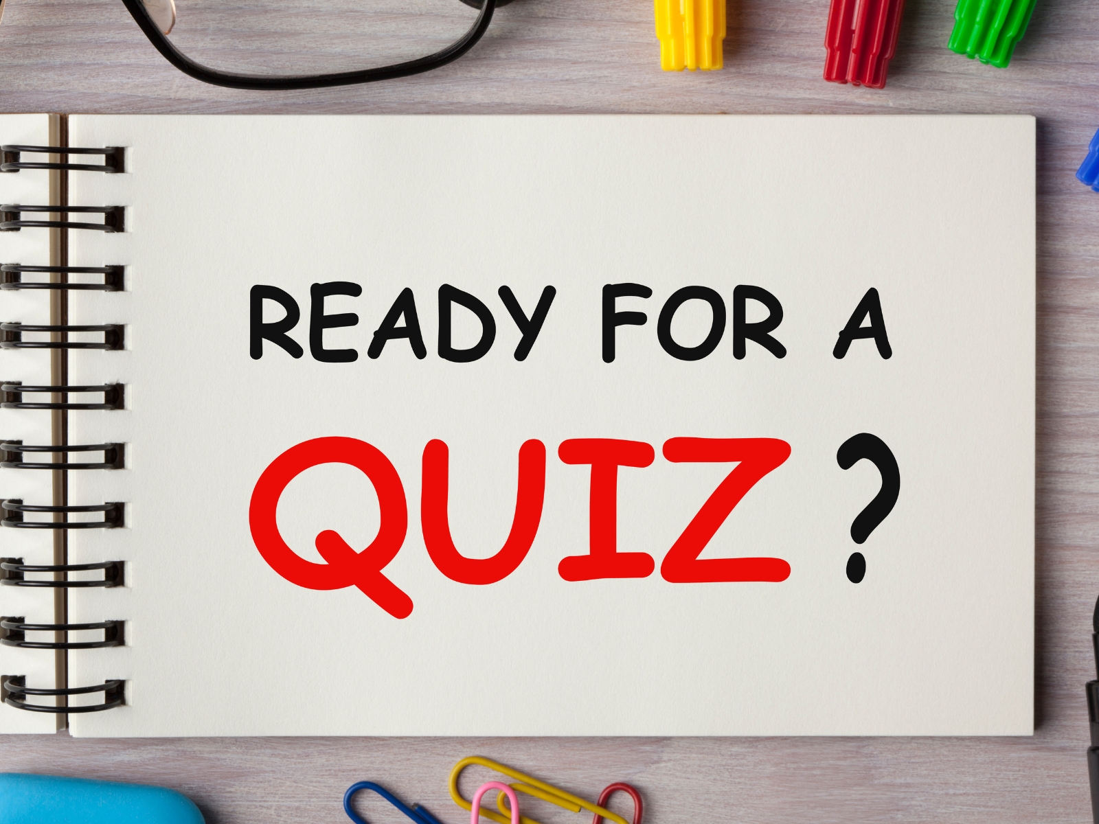 A spiral notebook on a desk with the text "Ready for a QUIZ?" written on its open page. The words "ready for a" are in black, and "QUIZ?" is in bold red. Colored markers, eyeglasses, and paperclips are scattered around the notebook.