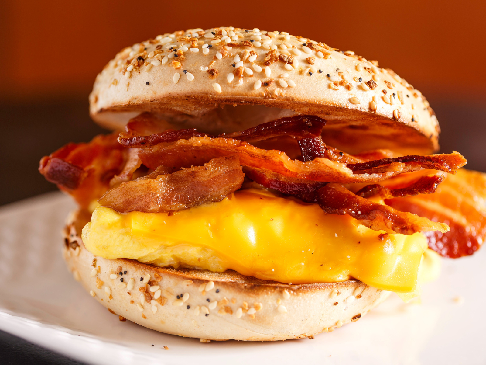 A close-up of a bagel sandwich featuring crispy bacon, a fluffy egg patty, and melted cheddar cheese, all nestled within a sesame and poppy seed bagel. The sandwich is placed on a white plate with a blurred, warm-toned background.