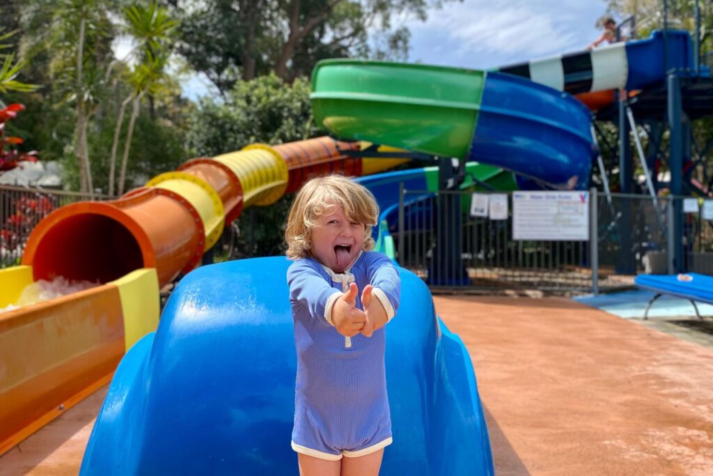 A young child with blond hair, wearing a blue swimsuit, stands at the bottom of a blue waterslide at Nambucca Heads, smiling and giving two thumbs up. Behind the child, a colourful setup with various slides is visible under a clear sky with trees in the background.