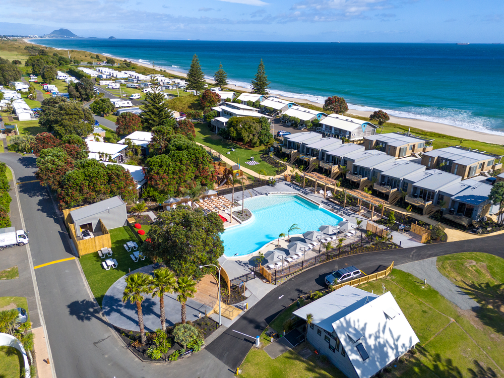 Aerial view of Tasman Holiday Parks - New Zealand featuring a large swimming pool surrounded by lounge chairs. The park has cabins and camping sites scattered among trees, with a beach and ocean in the background. Paths and roads weave through the landscape, offering scenic walks that connect various areas.