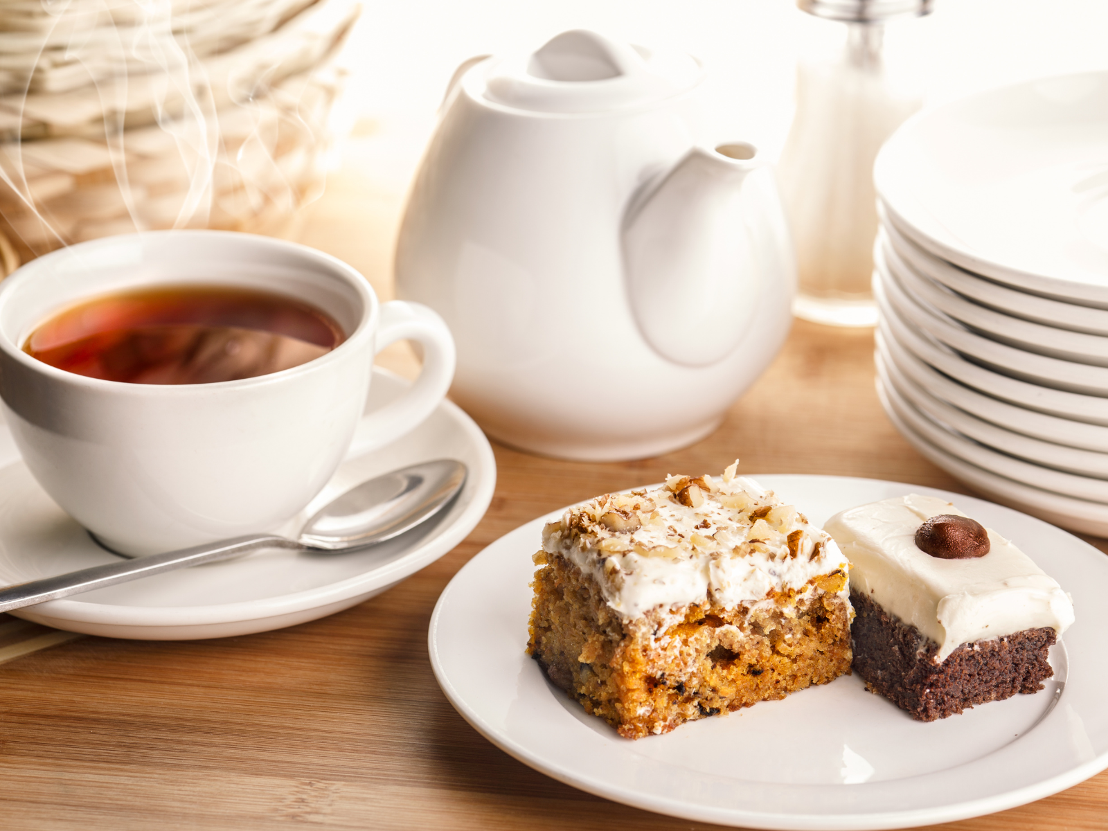 A close-up view of a white cup filled with tea next to a white teapot, a stack of white plates, and two pieces of cake on a white plate. One cake has a brown base with white frosting topped with a nut, and the other has a light brown base with white frosting.