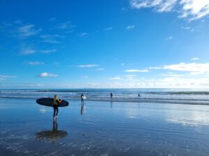 A person carrying a surfboard walks along the wet sand of a beach towards the waves, with three others already in the water. The sky is bright blue with scattered clouds and the sunlight reflects off the wet sand, creating a serene and peaceful scene.