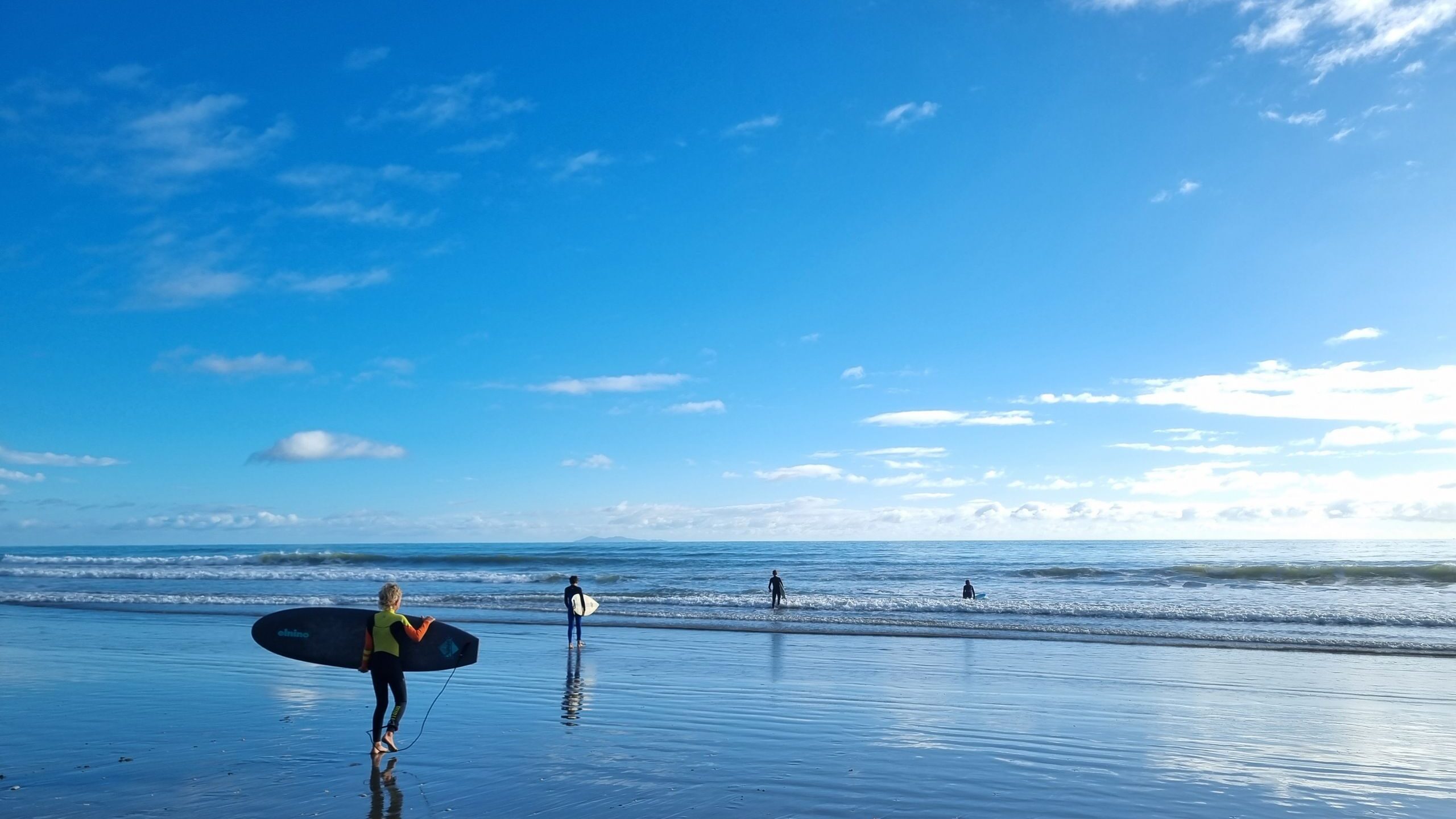 A person carrying a surfboard walks along the wet sand of a beach towards the waves, with three others already in the water. The sky is bright blue with scattered clouds and the sunlight reflects off the wet sand, creating a serene and peaceful scene.