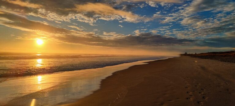 Papamoa Beach at sunrise with gentle waves washing onto the shore. The sky is a mix of vibrant blue and golden hues from the rising sun, partially obscured by scattered clouds. Footprints trail along the smooth, wet sand toward the horizon, where someone walks leisurely.