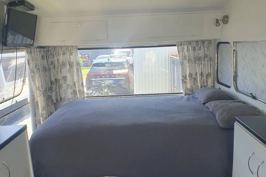 Interior of a cozy camper van bedroom featuring a neatly made bed with a quilt and two pillows. The room includes built-in white cabinets on both sides and a flat-screen TV mounted above. A large rear window displays an exterior view with a parked car outside.