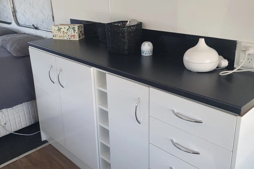 A sleek, modern white cabinet with a black countertop stands against a wall. It has four drawers, two doors, and a central wine rack. Items on the counter include a mirror, black basket, small decorative house, spherical light, and white essential oil diffuser.