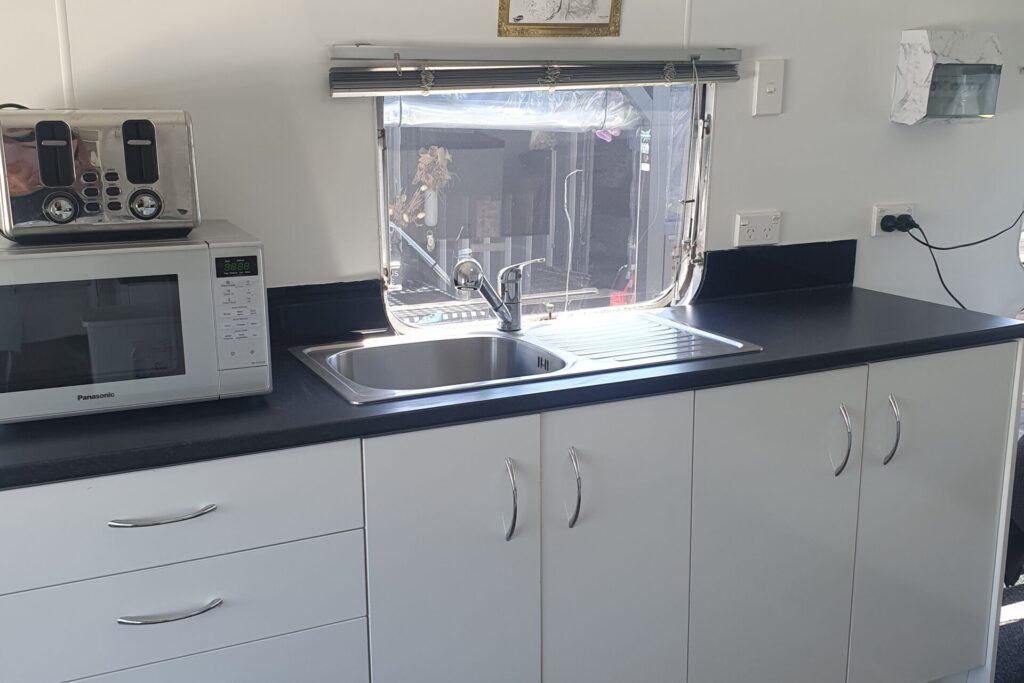 A small, modern kitchen with white cabinets and a black countertop. A stainless steel sink is centered below a window. Above the sink is a framed drawing. On the left side of the counter, there are two microwaves stacked. Kitchen utensils are stored on the right.