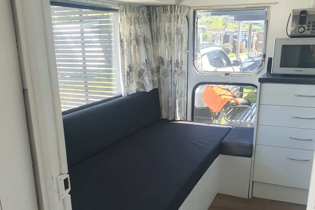 The image shows the interior of a small camper or RV. There is a built-in black bench seat with storage underneath, a window with blinds and floral curtains, a counter with drawers, and a microwave on top. The space is bright with natural light from the windows.