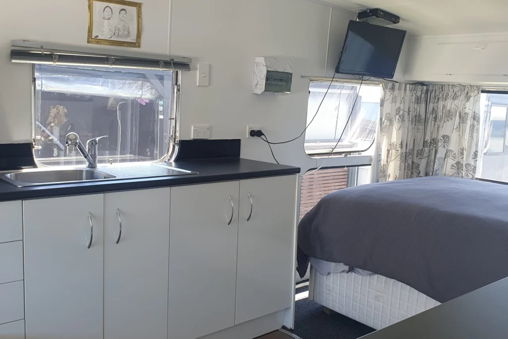 Interior of a small, modern RV with white cabinetry and black countertops. A bed with gray bedding is situated at the rear. The kitchen area features a sink, and a small TV is mounted on the wall. A large window with patterned curtains provides natural light.