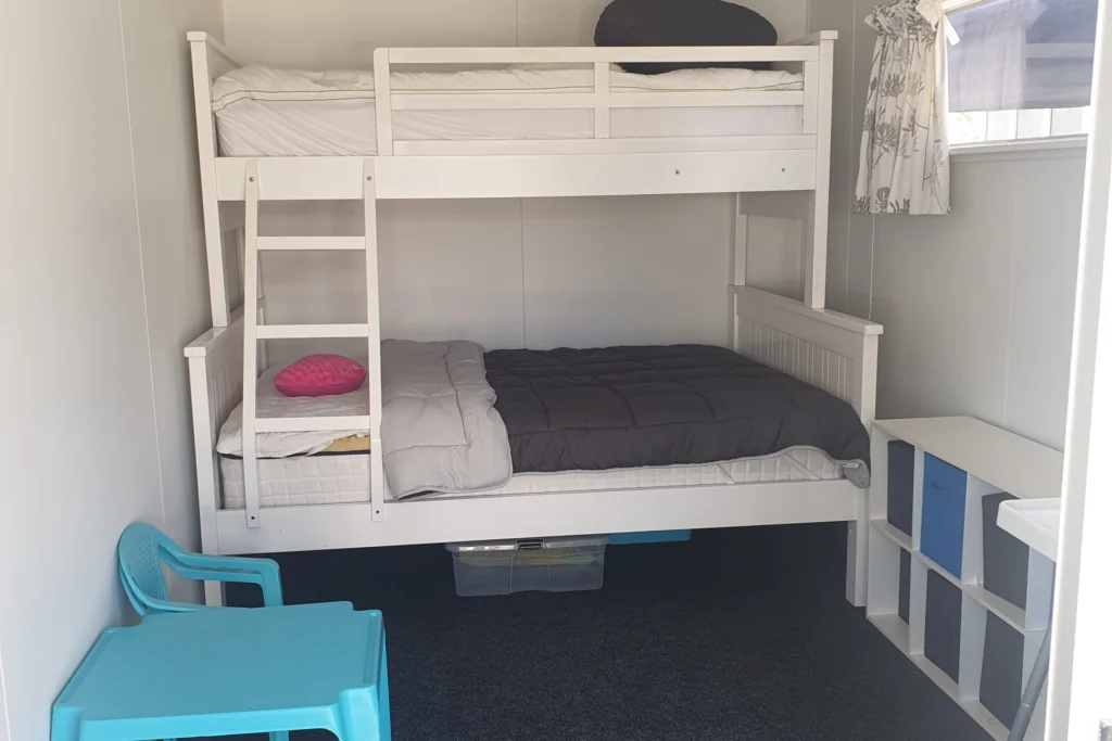 A small room with white walls features a bunk bed; the lower bunk is double-sized with a gray comforter and pink pillow, and the upper is single-sized with a white comforter and black pillow. A turquoise chair sits next to the bed. A small window with a curtain is on the right.