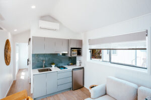 A modern, minimalist kitchen and living area inspired by Airlie Beach. The kitchen features gray cabinets, a sink, a stovetop, a microwave, and a small refrigerator. The living area has a light-colored armchair next to a large window with a roller shade and a wall-mounted air conditioner.