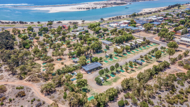 Aerial view of a coastal caravan park with rows of caravans and trailer sites arranged on a grid-like pattern amidst trees. A few buildings and amenities are scattered within the park. The coast with a sandy beach and ocean is visible in the background.
