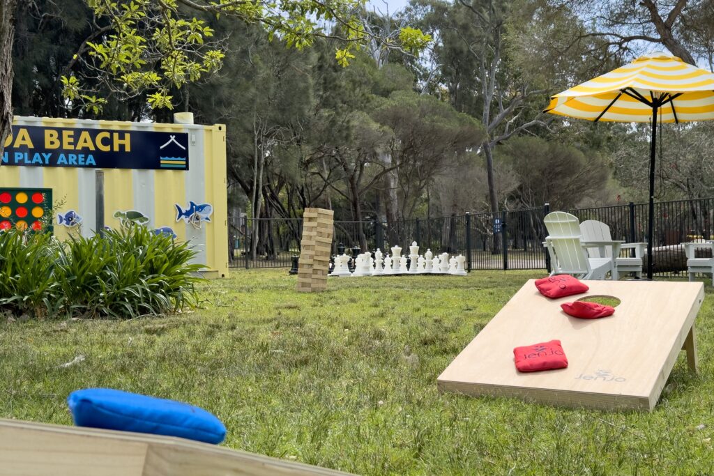 A grassy play area near Kioloa Beach features large outdoor games, including a Jenga tower, a Connect Four board, and a chess set. Colorful bean bags are set up on a cornhole board. In the background, there's a yellow-striped umbrella and chairs.